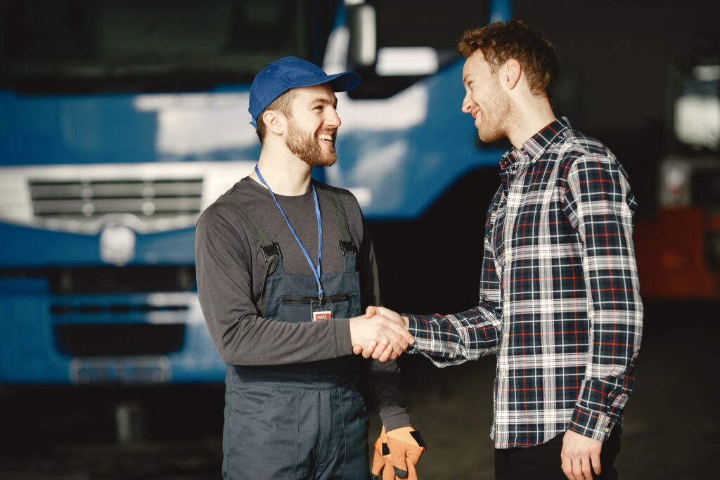 project-3 Mechanic in uniform and customer in plaid shirt shaking hands in front of a blue truck inside a garage.